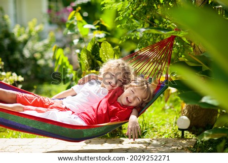 Kids relax in colorful rainbow hammock. Hot day garden outdoor fun. Boy playing outdoor. Brothers play outside in sunny backyard. Afternoon nap during summer vacation. Children relaxing.