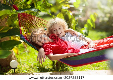 Kids relax in colorful rainbow hammock. Hot day garden outdoor fun. Boy playing outdoor. Brothers play outside in sunny backyard. Afternoon nap during summer vacation. Children relaxing.