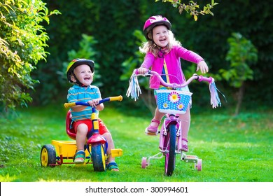 Kids Riding Bikes In A Park. Children Enjoy Bike Ride In The Garden. Girl On A Bicycle And Little Boy On A Tricycle In Safety Helmet Playing Together Outdoors. Preschool Child And Toddler Kid Biking.