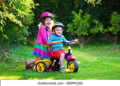 Kids Riding Bikes In A Park. Children Enjoy Bike Ride In The Garden. Girl On A Bicycle And Little Boy On A Tricycle In Safety Helmet Playing Together Outdoors. Preschool Child And Toddler Kid Biking.