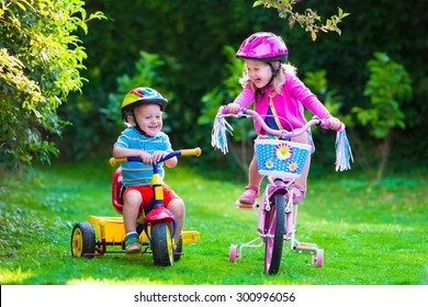 Kids Riding Bikes In A Park. Children Enjoy Bike Ride In The Garden. Girl On A Bicycle And Little Boy On A Tricycle In Safety Helmet Playing Together Outdoors. Preschool Child And Toddler Kid Biking.