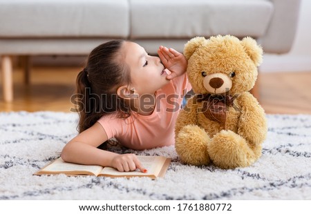 Kid's Secrets. Little Asian Girl Reading Book Whispering Sharing Secret With Teddy Bear Playing Lying On Floor At Home.