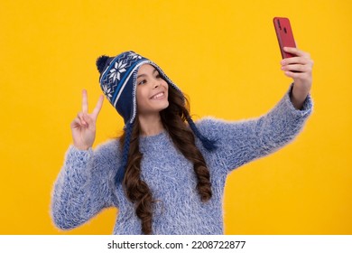 Kids Selfie. Close-up Portrait Of Cute Teen Girl In Winter Knitted Hat Using Mobile Phone, Cell Web App, Isolated Over Yellow Color Background. Happy Face, Positive And Smiling.