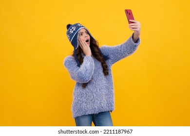 Kids Selfie. Close-up Portrait Of Cute Teen Girl In Winter Knitted Hat Using Mobile Phone, Cell Web App, Isolated Over Yellow Color Background. Surprised Teenager Girl.