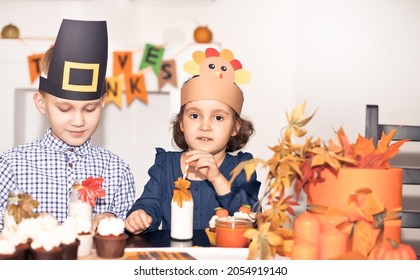 Kids Sitting On Festive Table And Celebrating Thanksgiving Day. Children In Paper Turkey Hat And Pilgrim Hats Eating Cupcakes And Drinking Milk.