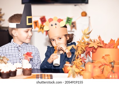 Kids Sitting On Festive Table And Celebrating Thanksgiving Day. Children In Paper Turkey Hat And Pilgrim Hats Eating Cupcakes And Drinking Milk.