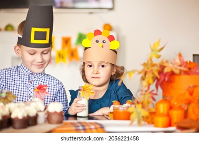 Kids Sitting On Festive Table And Celebrating Thanksgiving Day. Children In Paper Turkey Hat And Pilgrim Hats Eating Cupcakes And Drinking Milk.