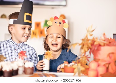 Kids Sitting On Festive Table And Celebrating Thanksgiving Day. Children In Paper Turkey Hat And Pilgrim Hats Eating Cupcakes And Drinking Milk.