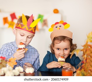 Kids Sitting On Festive Table And Celebrating Thanksgiving Day. Children In Paper Turkey Hat And Pilgrim Hats Eating Cupcakes And Drinking Milk.