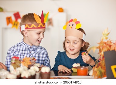 Kids Sitting On Festive Table And Celebrating Thanksgiving Day. Children In Paper Turkey Hat And Pilgrim Hats Eating Cupcakes And Drinking Milk.