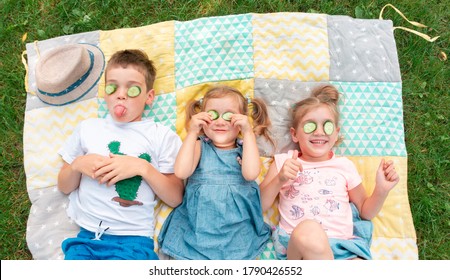 Kids Smile And Indulge Lying On A Patchwork Rug. Children Fooling Around With A Cucumber Mask. The Boy Shows His Tongue. Family Summer Camp Activity Ideas. Happy Kids Spend Their Summer Holidays. 