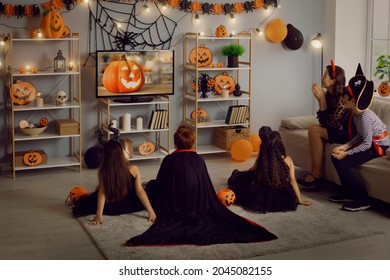 Kids In Spooky Costumes Of Witches, Monsters And Pirates Watching Children's Halloween Movie Sitting On Floor And Sofa In Interior Decorated With Orange And Black Pumpkins At Fancy Dress Party At Home