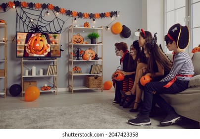 Kids In Spooky Costumes Of Witches, Pirates And Vampires Together Watching Children's Halloween Movie Sitting On Sofa In Interior With Festive Orange Black Pumpkin Decor At Fancy Dress Party At Home