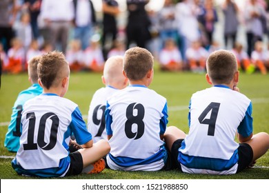 Kids Sports Team On Football Field. Group Of Children Sitting On Grass Soccer Pitch. Boys Wearing White Soccer Jersey Shirts With Black Numbers On Back