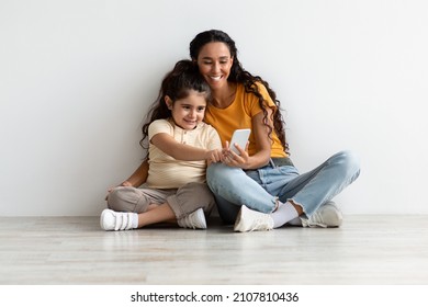 Kids And Technologies. Cheerful Little Girl Using Smartphone While Relaxing With Mom At Home, Happy Middle Eastern Mother And Daughter Browsing New Application On Mobile Phone And Smiling, Copy Space