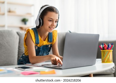 Kids And Technology Concept. Portrait of smiling girl sitting at table and using laptop computer, wearing wireless headphones, watching online course or movie, playing online game, free copy space - Shutterstock ID 2026218206