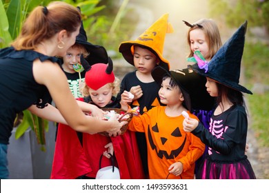 Kids trick or treat on Halloween night. Mixed race Asian and Caucasian children at decorated house door. Boy and girl in witch and vampire costume and hat with candy bucket and pumpkin lantern.  - Shutterstock ID 1493593631