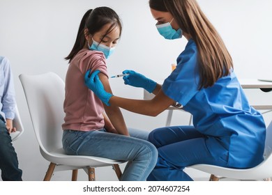 Kids Vaccination Against Covid-19. Asian Girl Getting Vaccinated Against Coronavirus In Modern Clinic. Nurse Wearing Protective Face Mask Making Antiviral Vaccine Injection Vaccinating Child