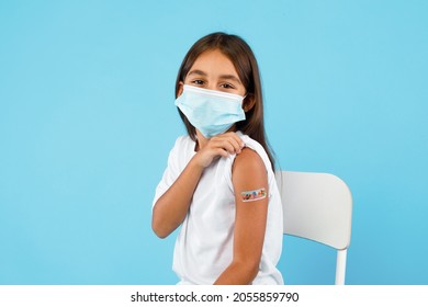 Kids Vaccination. Vaccinated Arabic Little Girl Showing Arm With Plaster After Vaccine Shot Looking At Camera Wearing Face Mask Over Blue Background. Studio Shot. Covid-19 Protection For Child