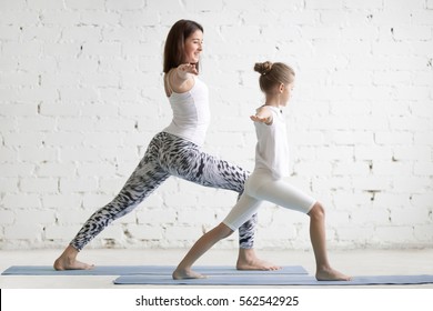 Kids yoga female teacher training a little child, practicing yoga, standing in Warrior one exercise, Virabhadrasana I pose, working out wearing sportswear, indoor full length, white studio background 