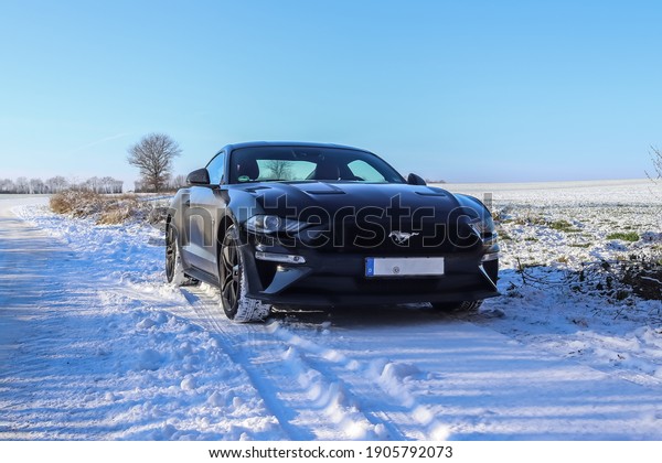 Kiel,\
Germany - January 30th 2021: Black Ford Mustang sports car in front\
of a winter landscape in Germany on a sunny\
day