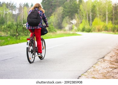 Kiev, Ukraine. May 1, 2020. A Girl, Young Woman In Checkered Shirt, Red Pants, Black Backpack Riding Bicycle On A Country Paved Road In Woods, Park In Spring, Summer. Journey Alone, Healthy Lifestyle.