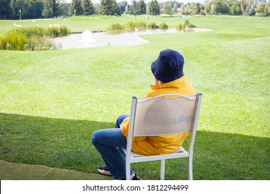 KIEV, UKRAINE - SEPTEMBER 10, 2020: A Woman In A Bright Yellow Jacket And A Panama Hat Is Sitting On A Green Golf Course After A Game On A Sunny Day. Back View