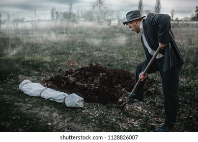 A man wearing a suit and hat is digging in the soil. The landscape looks very desolate.