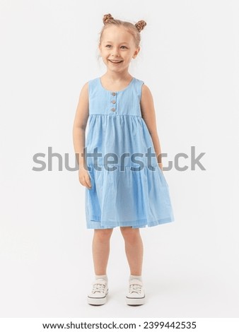 A kind 5-year-old girl with a beautiful smile with a fashionable hairstyle stands on a white background in a blue summer sundress and laughs.