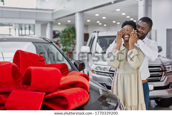 kind black man\
gave a car to his wife, the car is wrapped in a red bow, man closed\
woman\'s eyes, prepared\
surprise