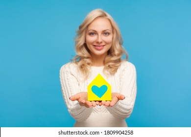 Kind Blond Woman Holding In Hands Paper House Model With Painted Heart, Charity, Orphanage And Shelter For People In Need. Indoor Studio Shot Isolated On Blue Background