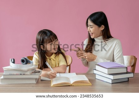 A kind and caring Asian female teacher or sister helps a young, adorable girl do homework at a study table against an isolated pink background. Education concept