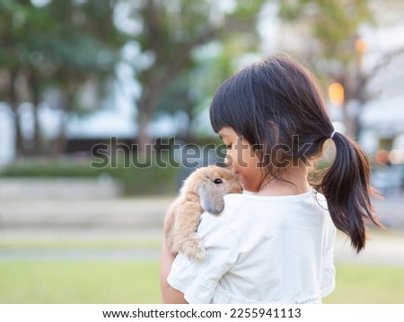 Kind little cute girl 7 years old holding a baby holland lop rabbit on shoulder at the garden. Child with a gentle heart, loves animals.