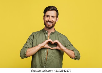 Kind Man Shaping Hands Like Heart, Making Love Gesture Near Chest, Expressing Kindness And Smiling At Camera, Standing Over Yellow Studio Background