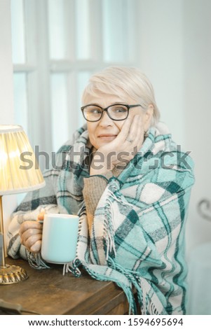 Kind Old Granny Thinking About Something And Drinking Tea. The Thoughtful Woman Cups Chin In Her Hand. She Is Relaxing At Home With A Cup Of Tea In Her Hand.