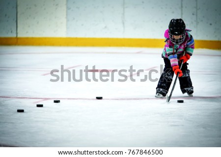 A kindergarten aged little girl in ski pants and winter jacket and helmet lining up a row of hockey pucks to practice shooting at a net