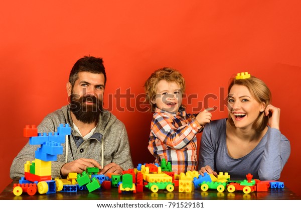 Kindergarten and family concept. Family with
cheerful faces build toy cars out of colored construction blocks.
Kid points at toy on moms head. Man with beard, woman and boy play
on red background.