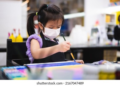 Kindergarten Girl Is Doing Water Color Painting In Classroom. Children Wear White Face Mask While Sitting In Special Class, To Prevent Spread Of Virus And Dangerous PM2.5 Particulate Matter For Health