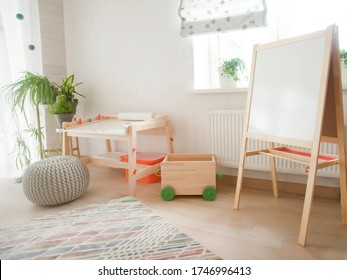 Kindergarten Room With Easel Chair And Table For Painting. Children  Space. Empty White Desk At Kindergarten. No People. Modern Interior. Sunlight And Plants On Windowsill