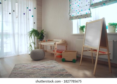 Kindergarten Room With Easel Chair And Table For Painting.  Empty White Desk At Kindergarten. No People. Modern Interior. Sunlight And Plants On Windowsill. Children  Space.