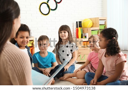 Kindergarten teacher reading book to cute little children indoors