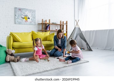 Kindergarten Teacher Sitting On Carpet And Playing With Toddler Girl With Down Syndrome Near Child In Playroom 