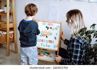 Kindergartener stand with back and learn english letters, alphabet and numbers on magnetic board. Educator sit near boy in playroom and check exercise. Reading and counting, study in daycare centre. 