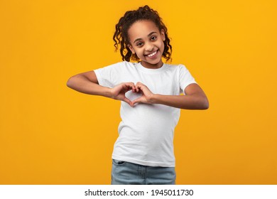 Kindness Concept. Portrait of cute African Amercian girl making heart shape with her hands and fingers near chest, smiling to camera, posing over yellow background in studio, copy space - Powered by Shutterstock