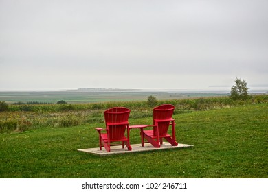 Kings County, Nova Scotia, Canada: Two Red Adirondacks Chairs Look Toward The Minas Basin At The Grand-Pré National Historic Site, On A Cloudy Summer Day.