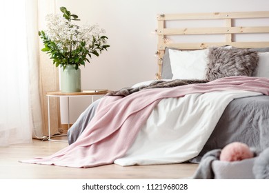 King-size bed with grey sheets and pink blanket standing in bright bedroom interior with fresh plants in vase and window with drapes