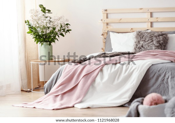 King-size bed with grey sheets and pink blanket\
standing in bright bedroom interior with fresh plants in vase and\
window with drapes