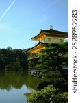 Kinkaku-ji, the Golden Pavilion, a famous Zen temple in Kyoto, Japan. The temple is reflected in a serene pond surrounded by lush greenery and a clear blue sky with fluffy clouds.