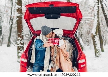Kissing couple in love sitting in car trunk holding cups with tea in knitted Christmas cases in snowy winter forest. Close up of hot drinks