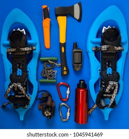 Kit Of Gear For Hiking, Adventure And Survival In Wilderness During Winter - Ax, Snowshoes, Gloves, Saw, Gps Receiver And Other Items Lie On A Blue Background. Flat Lay, Top View.